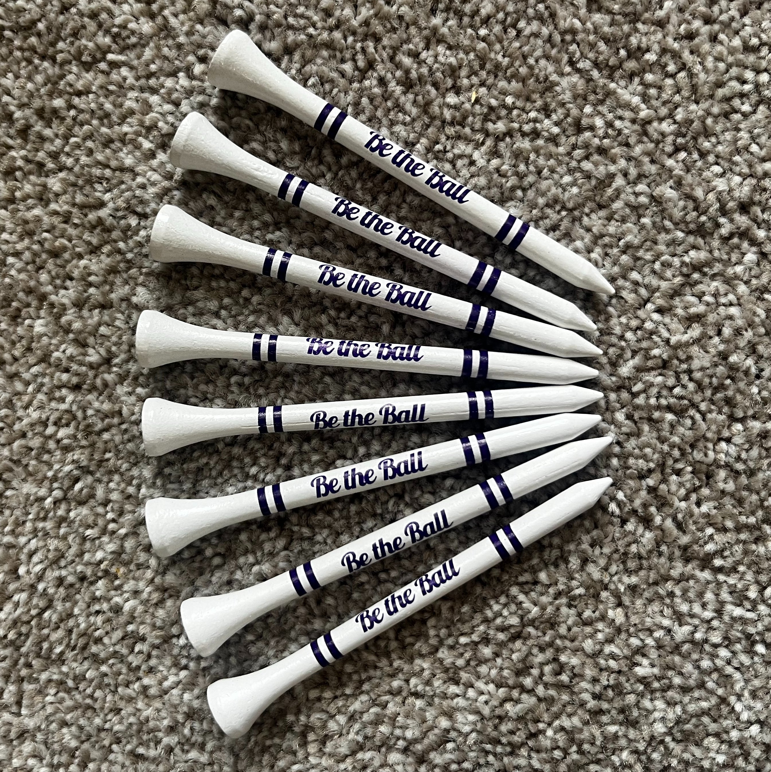 White golf tees with 'Be the Ball' text on a beige carpet