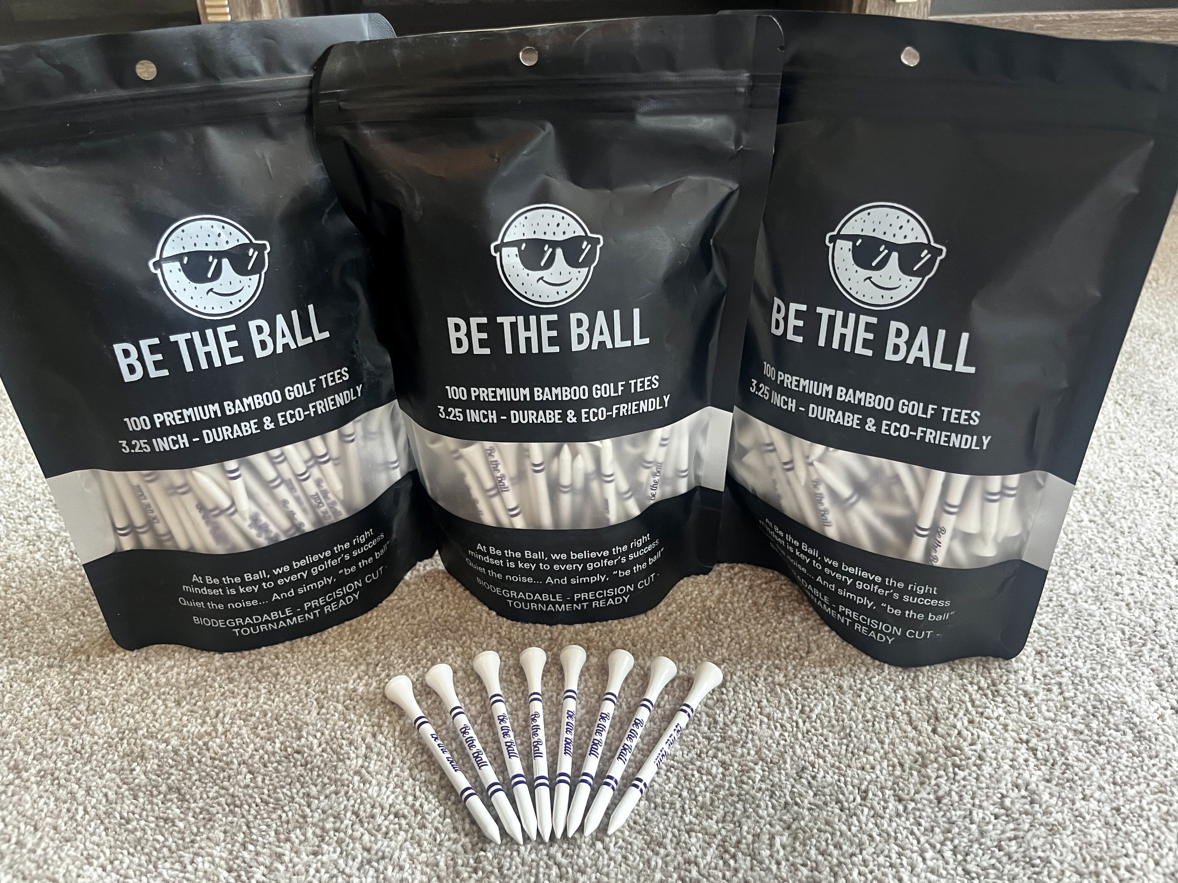 Three packages of 'Be The Ball' golf tees with individual tees displayed on a carpeted floor.